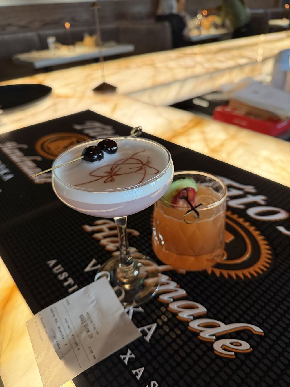 Two Kasi cocktails on the bar mat, a pale coupe with brandied cherries and a rocks pour garnished with fruit