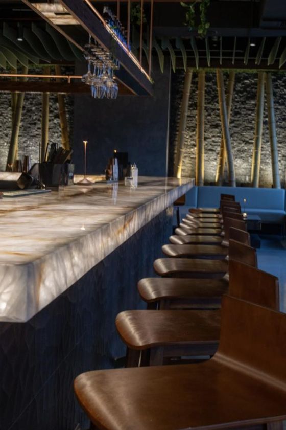 The Kasi bar seen down its length, glowing onyx counter, hanging glassware, and a lit bamboo wall behind