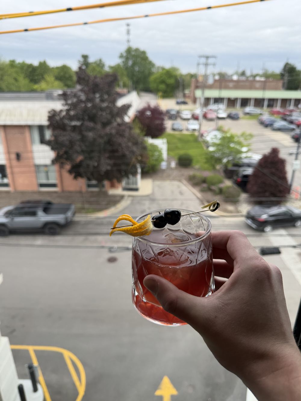 A deep red stirred cocktail with an orange twist and brandied cherries held up against a window