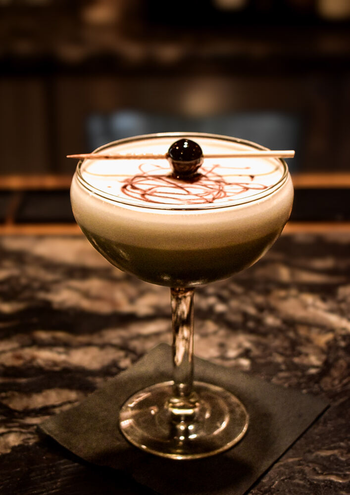 A dark cocktail served up in a coupe with a thick foam cap, bitters drawn across the top and a brandied cherry on a pick