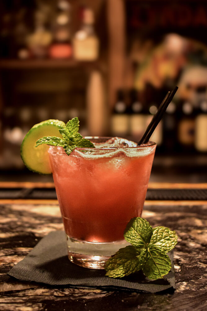 A tall red cocktail over ice garnished with a cucumber wheel and a sprig of mint, sat on a black napkin