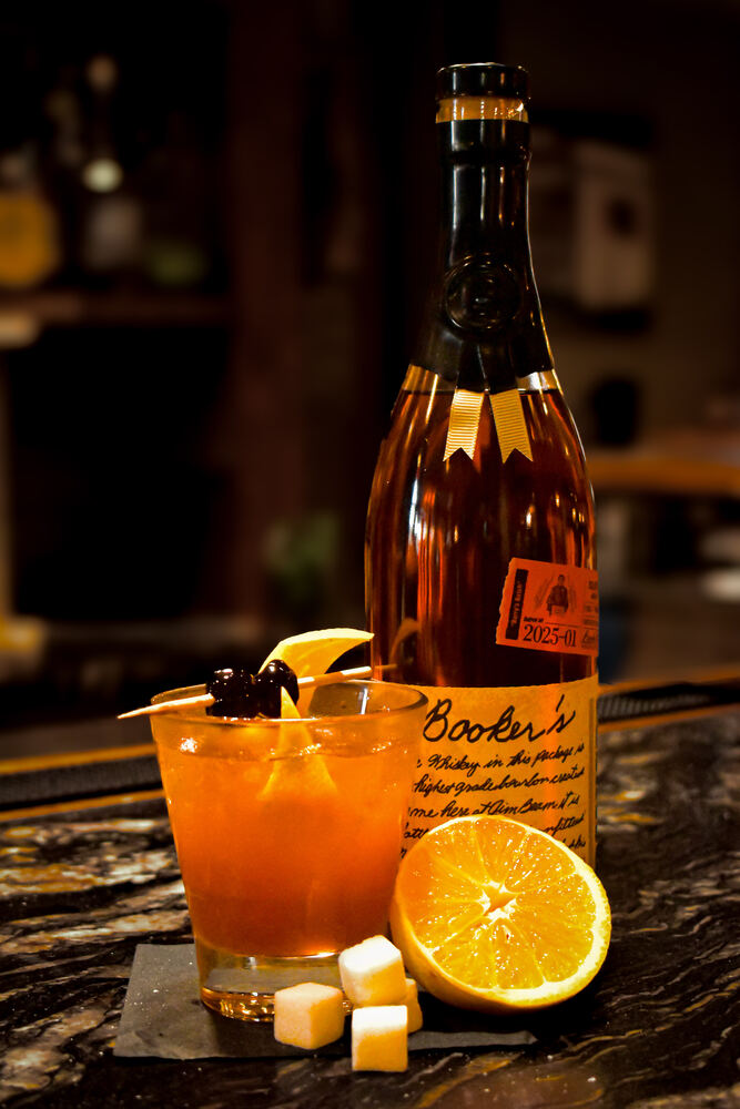 An old fashioned with an orange peel and brandied cherries next to a bottle of Booker's bourbon and sugar cubes