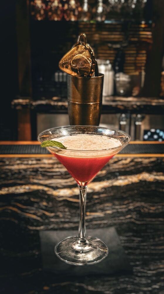 A red shaken cocktail in a martini glass with a mint leaf, staged in front of a shaker tin and strainer
